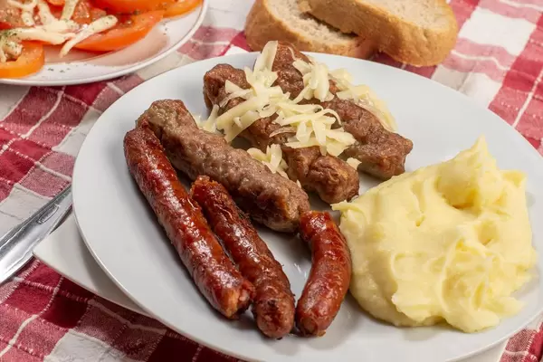Teller mit Cevapcici aus Hackfleisch bestreut mit Käse, dazu Kartoffelbrei, Brot und Tomaten