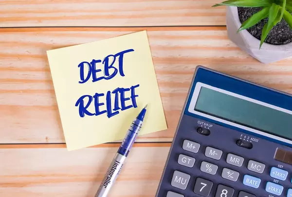 Text Debt Relief auf einer Haftnotiz mit Kugelschreiber, Taschenrechner und Tischpflanze auf einem Holztisch