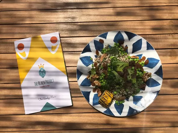 The Beach Project Sandy Menu next to a veggie bowl, with quinoa, wild greens, broccoli, green peas, walnuts, raisins, corn & parian bread on a wooden table