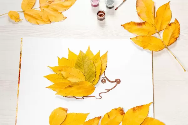 The hedgehog is made of autumn yellow leaves with foliage and paints on a white wooden table (Flip 2019)