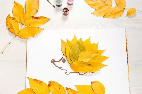 The-hedgehog-is-made-of-autumn-yellow-leaves-with-foliage-and-paints-on-a-white-wooden-table.jpg