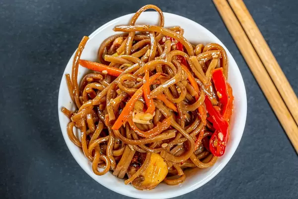 Thin buckwheat noodles with sauce, vegetables, mushrooms and sesame in bowl on black background with chopsticks