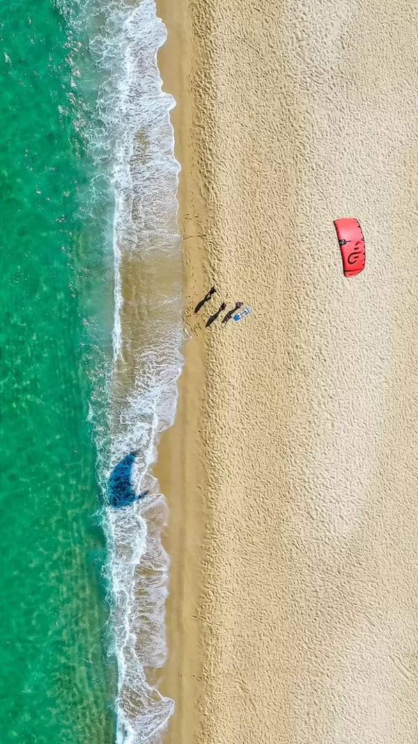 Three people fly a red kite on a sandy beach. Windy beach of Mikri Vigla, Naxos. Overhead drone shot