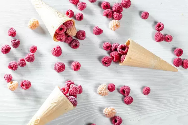 Three waffle cones with red and yellow raspberry on white wooden background
