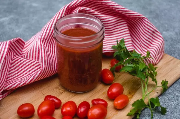 Tomatensoße in einem Aufbewahrungsglas umgeben von Tomaten und Petersilie auf einem Holzschneidebrett