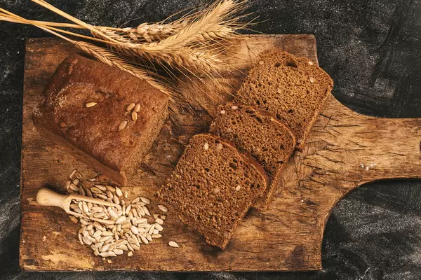 Top view, black bread with sunflower seeds in a wooden scoop and ears of wheat on an old cutting board