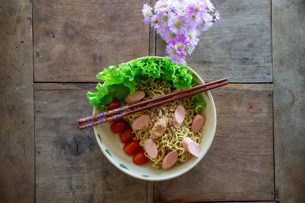 Top View Food Photo of Dried Instant Noodles with Sausages, Cherry Tomatoes, Peanuts and Lettuce on a Wooden Table with Flowers