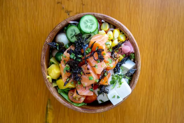 Top View Food Photo of Hawaiian Poke Bowl with fresh Vegetables, Tofu, Quail Eggs, Mango, dried Seaweed, fried Onions and Salmon in a Wooden Bowl