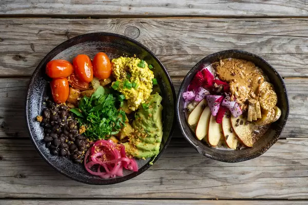 Top View Food Photo of Healthy Breakfast Porridge with Fresh Fruits and Peanut Butter and a Healthy Vegan Lunch with Black Beans, Avocado, Baked Potatoes and Cherry Tomatoes on a Wooden Table