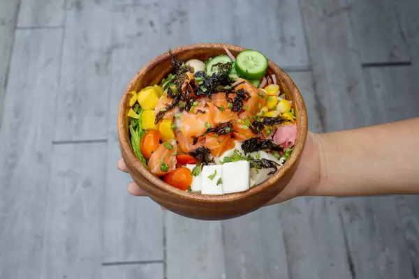 Top View Food Photo of Person holding a Wooden Bowl with Healthy Hawaiian Poke Bowl with Fresh Mango, Tofu, Vegetables and Raw Salmon