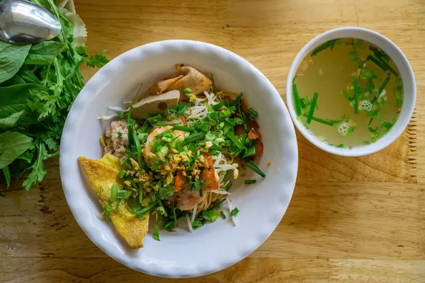 Top View Food Photo of Vietnamese Dry Hu Tieu Noodles with Pork, Shrimp, Herbs, Fried Onion and Soup on a Wooden Table in a Restaurant