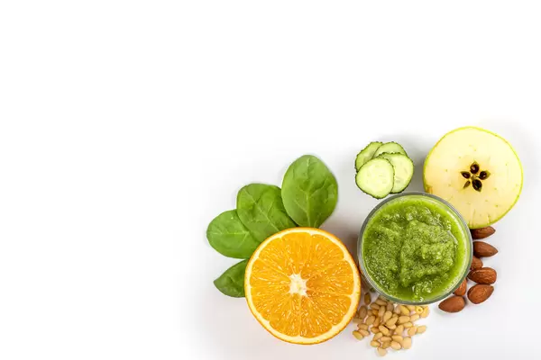 Top view, glass of green smoothie with halved apple, orange, chopped cucumber, almonds and pine nuts