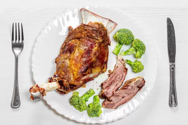Top-view Lammkeule mit Brokkoli auf einem weißen Teller mit Besteck und weißem Holztisch serviert