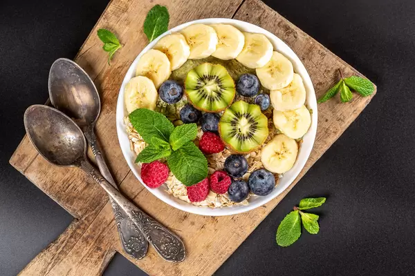 Top view, oatmeal with blueberries, raspberries, banana, kiwi, and mint on an old wooden kitchen board with two spoons