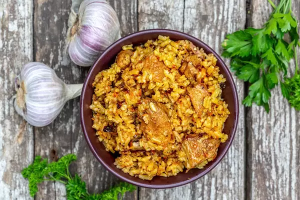 Top view of a bowl of meat pilaf on a wooden gray background with fresh parsley and garlic