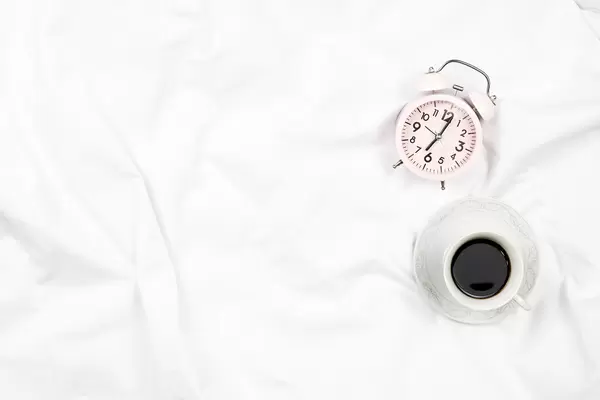 Top view of an alarm clock and a cup of black coffee on a cozy white bed