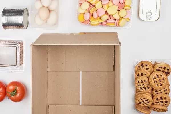 Top view of an empty cardboard box surrounded by food products: tomatoes, eggs, sweets, tin cans