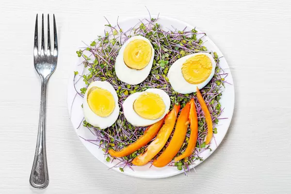 Top view of boiled eggs with micro greens and pieces of bell pepper in a plate on a white wooden background with a fork (Flip 2019)