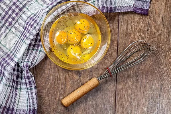 Top view of broken chicken eggs in bowl with whisk on wooden background