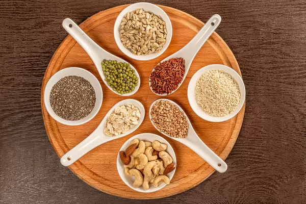 Top view of nuts and seeds in bowls and grains of cereals in spoons on round wooden board