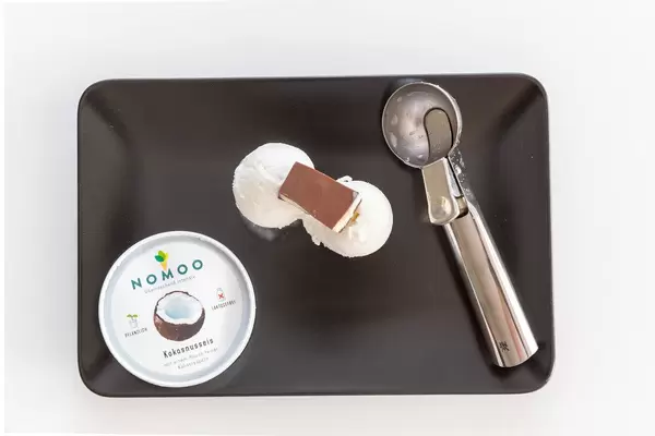 Top View of organic and vegan Nomoo Coconut ice cream and rasps with two scoops, topped with chocolate and ice cream spoon on a black plate