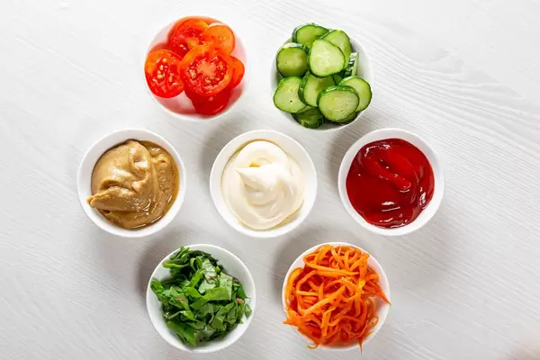 Top view of sauces, sliced vegetables and greens on white wooden background
