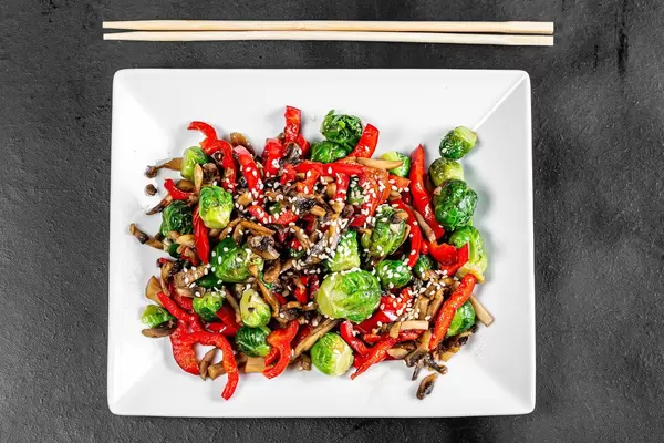 Top view of stewed vegetables and mushrooms with sesame seeds on black background with chopsticks