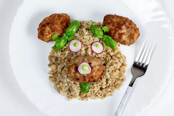 Top view of the face of a bear made of buckwheat porridge, meatballs, vegetables and basil on a white plate