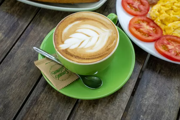Top View Photo of Cappuccino with Latte Art on a Saucer with Spoon and Sugar Sachet next to Scrambled Eggs with Tomatoes for Breakfast on a Wooden Table