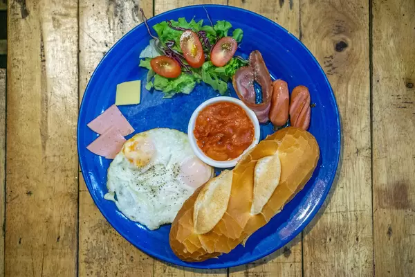 Top View Photo of Western Breakfast with Baguette, Sunny-Side Up Egg, Bacon, Sausage, Butter and Ham on a Blue Plate