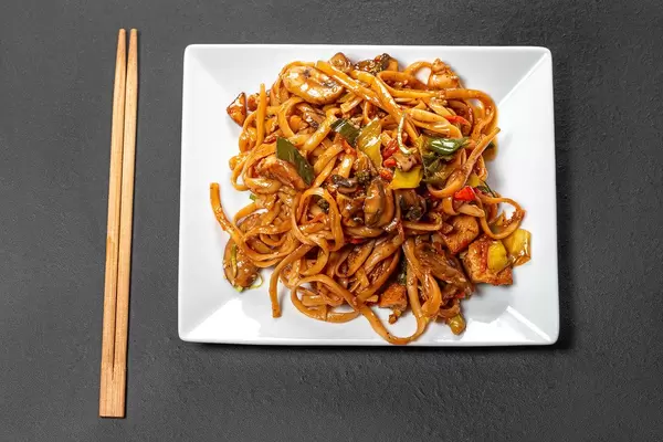Top view rice noodles with chicken, mushrooms and vegetables on a black background with chopsticks (Flip 2019)