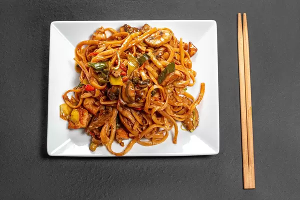 Top view rice noodles with chicken, mushrooms and vegetables on a black background with chopsticks