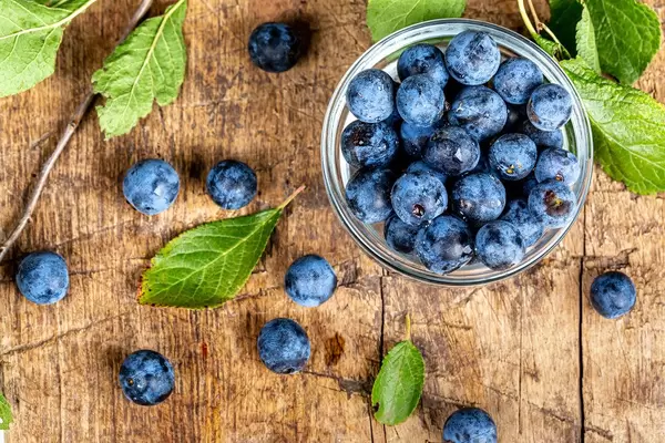Top view, ripe blackthorn berries in a glass bowl on a wooden background with green leaves