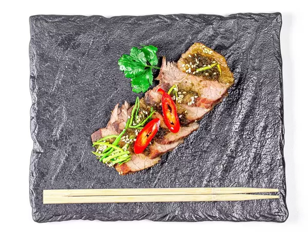 Top view, slices of baked beef with leek, hot chili pepper and sesame seeds on a black tray (Flip 2020)