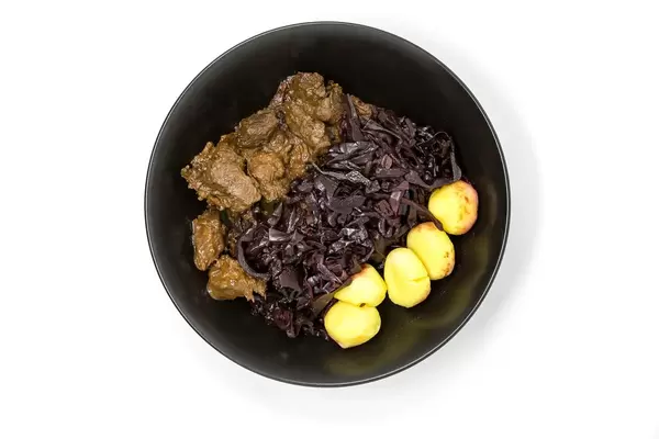 Traditionelles Rindergulasch mit Apfel-Rotkohl und Miniknödeln in einer schwarzen Schüssel - Aufsicht