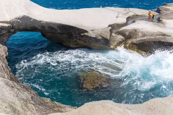 Two tourists walk on the rocks along the Aegean sea shores of Milos, among waves and natural arches