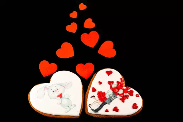 Two valentine gingerbread cookies with red small paper hearts on black background