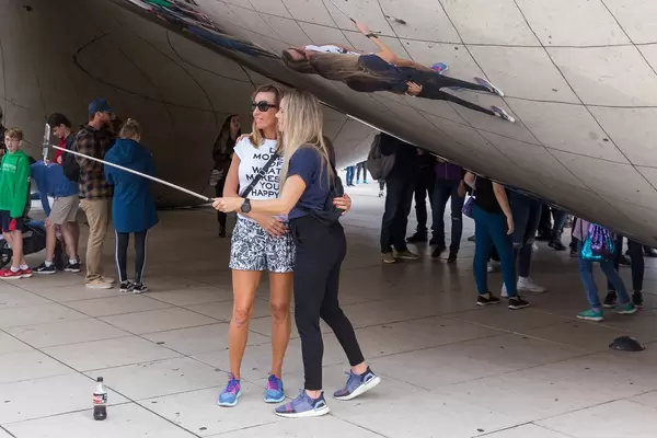 Two women take a photo with a selfie stick at the Cloud Gate public sculpture, AT&T Plaza, Millennium Park in Chicago, Illinois