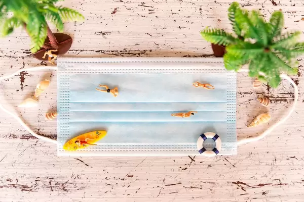 Urlauber im Sommer 2020 schützen sich vor Corona am Strand. Miniaturfiguren auf Mund-Nasen-Schutz