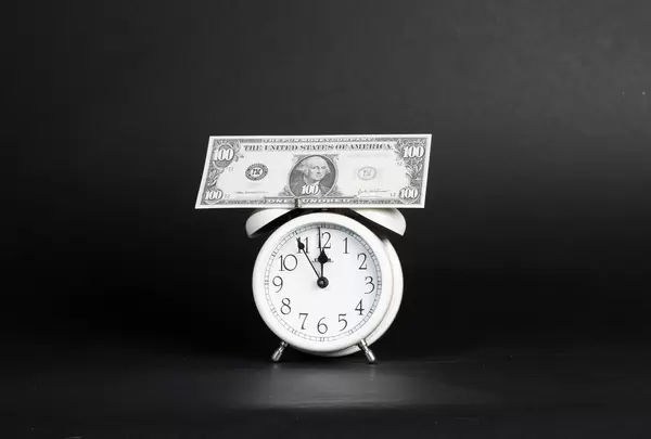 US-Dollar Banknote steht auf Wecker der auf 5 vor 12 steht vor dunklem Hintergrund