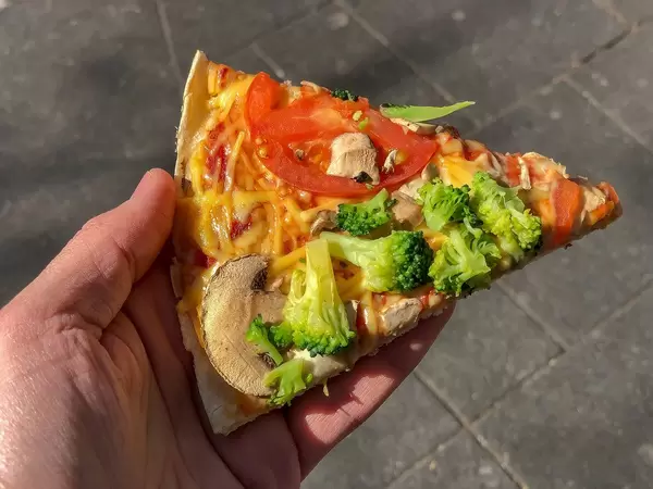 vegan Pizza slice "Kap Verde" from Dominos in Hand with Tomato, broccoli, fresh mushrooms and vegan processed cheese toppings