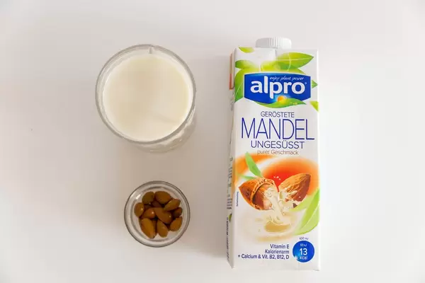 Vegane Milchverpackung von Alpro "Geröstete Mandel ungesüsst" liegt flach auf dem Tisch, neben einer Nussschale und einem laktosefreien Milchglas