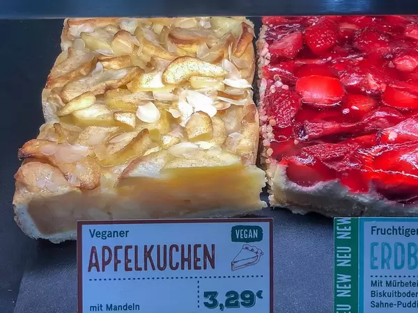 Veganer Apfelkuchen mit Mandeln und Preisschild, neben einem Stück Erdbeerkuchen in der Auslage