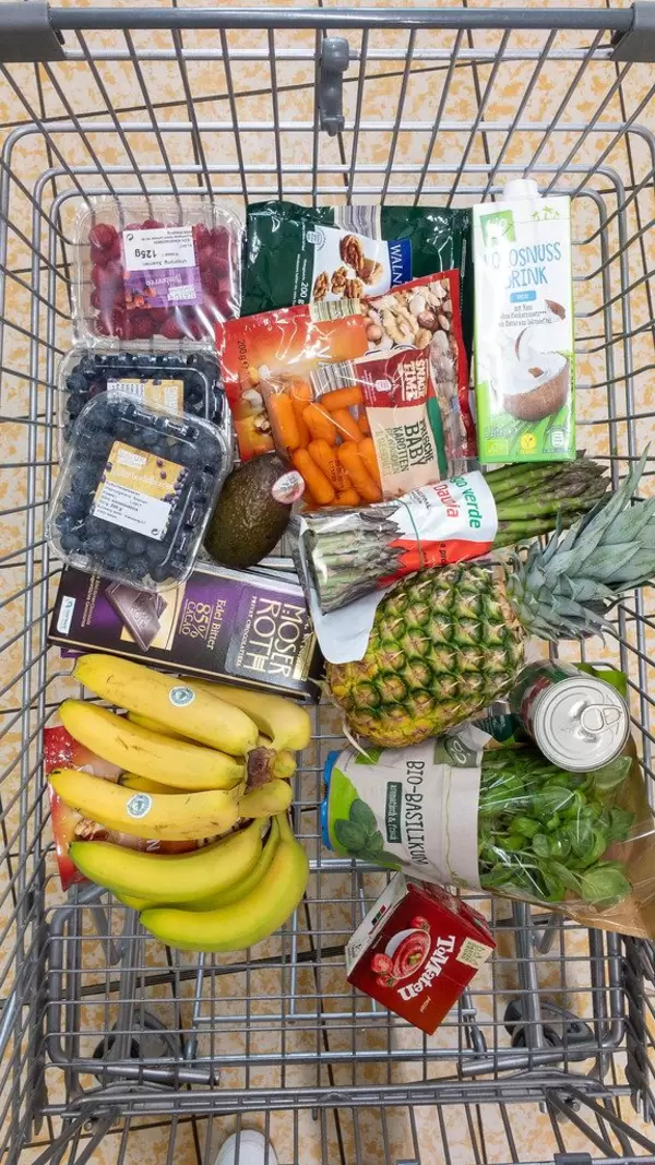 Veganer Einkauf im Einkaufswagen mit Beeren, Schokolade, Bananen, Ananas, frischem Basilikum, Baby-Karotten, Spargel, Kokosmilch, und Nüssen