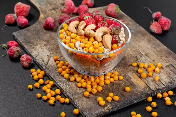 Veganes Frühstück in einer Glasschale mit Haferflocken, Erdbeeren, Cashew Nüssen und Sanddornbeeren auf einem Holzschneidebrett