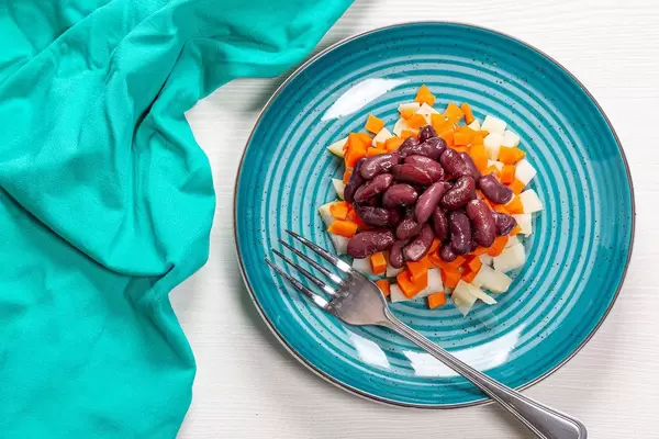 Vegetarischer, frischer Salat aus gekochten Kartoffeln, Karotten und Bohnen mit einer Gabel auf einem türkisen Teller und weißen Tisch