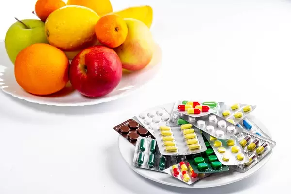 Viele bunte Tabletten und Medikamentenverpackungen vor einem Teller mit frischem, gesunden Obst
