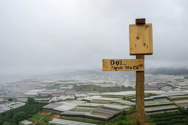 Vietnamese Words on Wooden Signboard with View of Grow Houses and Mountains in the Background