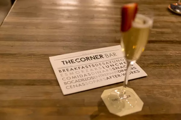 Visitenkarte der The Corner Bar wirbt mit Frühstück und Snacks, neben einem verschwommenen Sektglas mit Erdbeerscheibe, in Barcelona (Spanien)