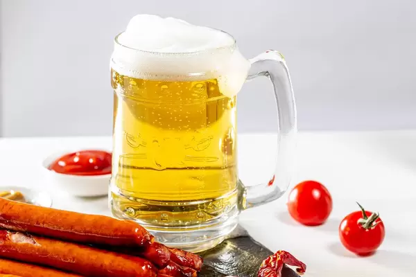 Volles Bierglas mit Schaumkrone zu knackigen Würstchen mit Ketchup und Senf vor weißem Tisch